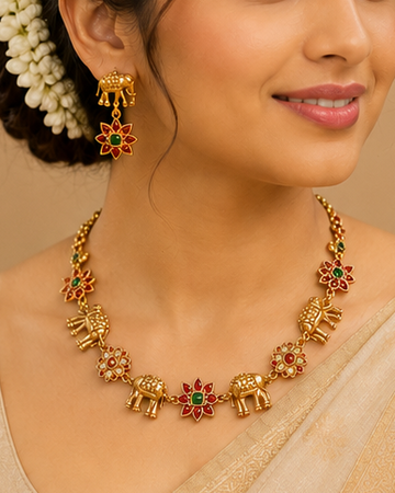 A woman wearing a beige saree and matching blouse, styled with a gold necklace and earrings, set against a warm traditional background with brass lamps.