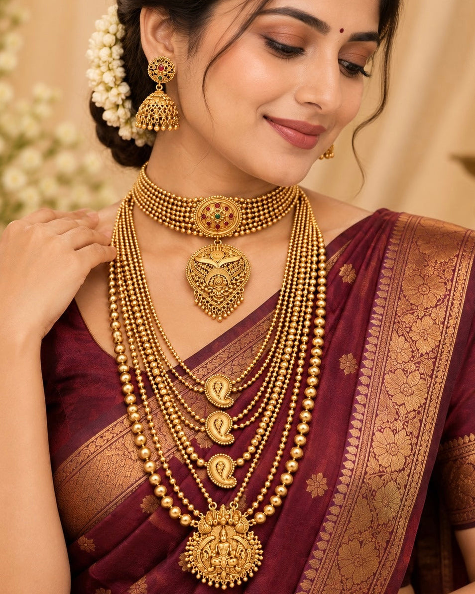 A woman wearing an ornate layered gold necklace set with a large pendant, matching earrings, and a deep purple saree with gold zari detailing.
