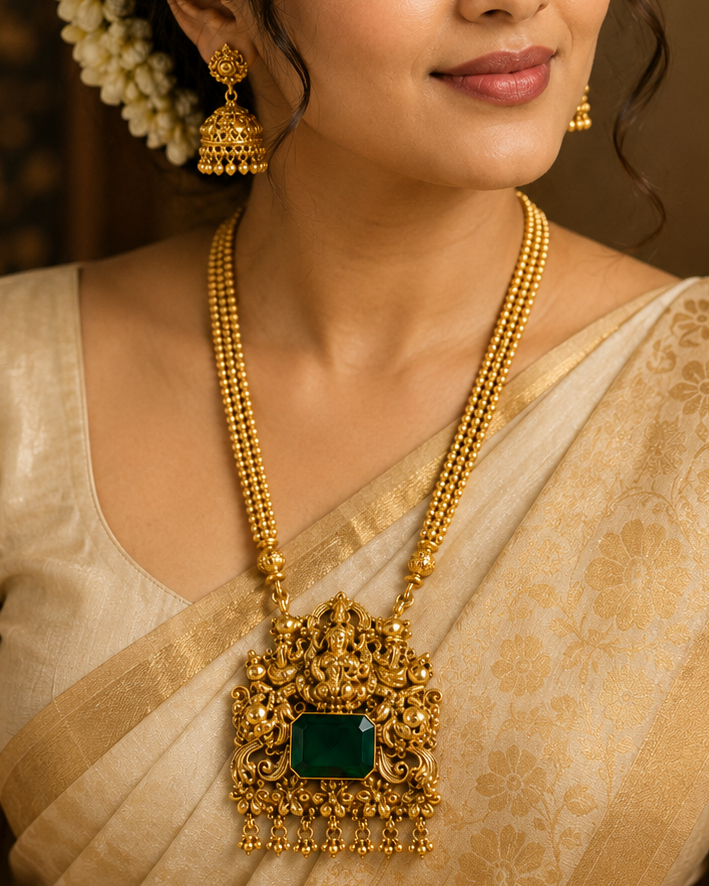 A woman wearing traditional Indian attire in a beige saree, styled with an ornate gold necklace featuring a large green stone pendant, matching gold earrings, and a white floral hair accessory.