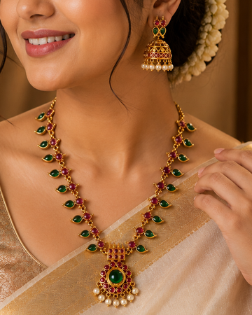 A woman wearing a traditional Indian outfit with a gold and cream saree, styled with a gold and green necklace and matching earrings, set against a warm, golden background.
