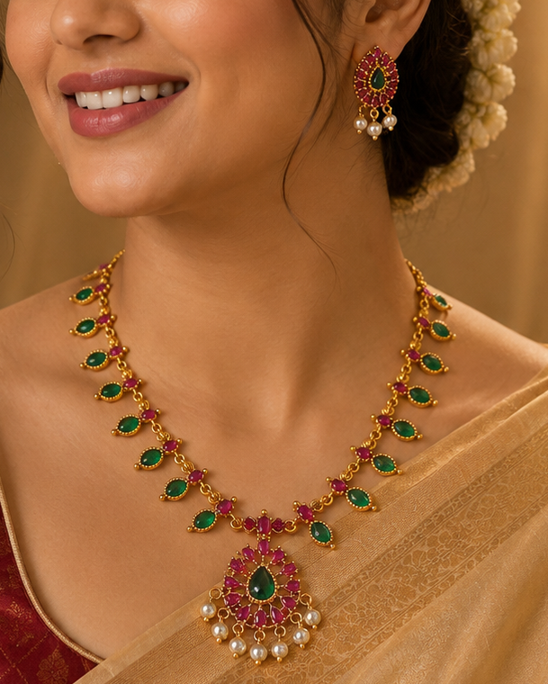 A woman wearing a gold-toned traditional necklace with green and red stones and matching earrings, styled with a cream flower in her hair and a beige saree with a maroon blouse.