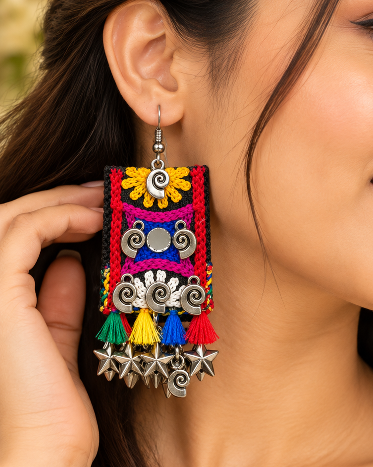 A close-up of a woman wearing colorful traditional Indian jhumka earrings with silver-toned metalwork, embroidered details, and hanging tassels in yellow, red, blue, and green.