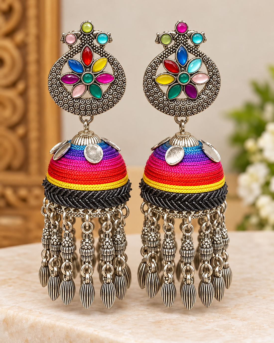 A woman wearing a pair of ornate Indian jhumka earrings with colorful embroidered details and dangling silver-toned chains, shown in a close-up portrait.