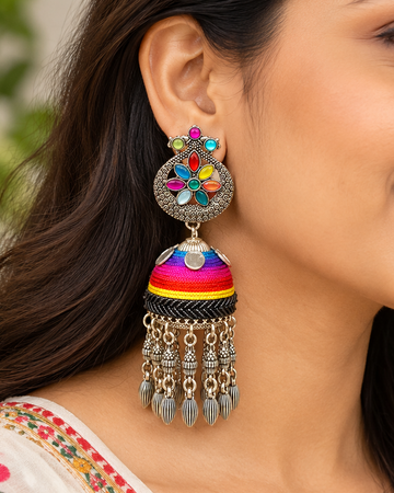 A woman wearing a pair of ornate Indian jhumka earrings with colorful embroidered details and dangling silver-toned chains, shown in a close-up portrait.