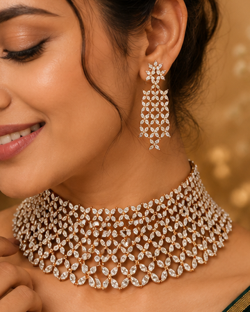 A woman wearing a sparkling gold-toned choker necklace and matching drop earrings, styled with a dark green velvet saree against a warm, elegant background.