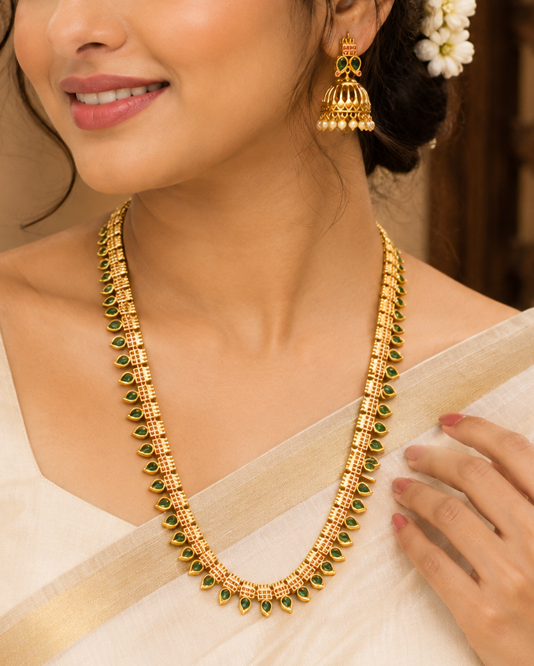 A woman wearing a gold necklace and matching gold earrings with green stone accents, styled with a white saree and white flowers in her hair.