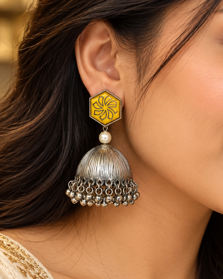 A close-up of a woman's ear wearing a traditional Indian jhumka earring. The earring features a yellow enamel hexagonal top with a black floral motif, a small white pearl, and a silver-toned bell-shaped dangle with hanging silver beads.