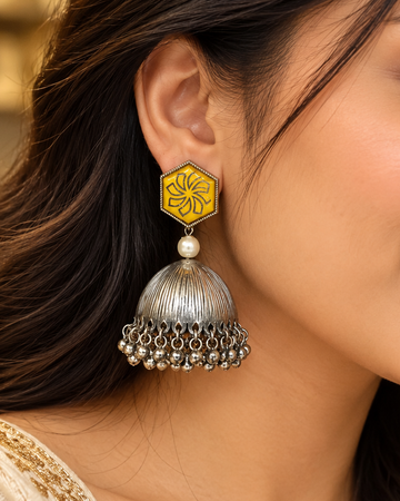 A close-up of a woman's ear wearing a traditional Indian jhumka earring. The earring features a yellow enamel hexagonal top with a black floral motif, a small white pearl, and a silver-toned bell-shaped dangle with hanging silver beads.