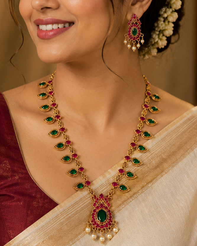 A woman wearing a traditional Indian outfit with a maroon blouse and cream saree, styled with a gold and red gemstone necklace and matching earrings.