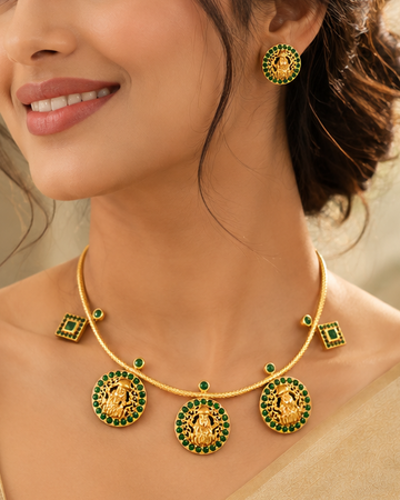 A woman wearing a gold and green traditional Indian jewelry set, including a necklace and matching earrings, styled with a beige saree.