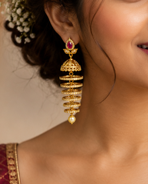 A close-up portrait of a woman wearing ornate gold dangling earrings with red and green stone accents, paired with a beige and maroon saree.