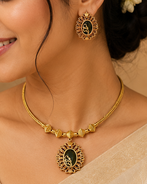 A woman wearing a gold-toned traditional necklace and matching earrings with dark oval centers, styled with a cream-colored sari and white hair flowers, in an indoor setting with warm lighting.