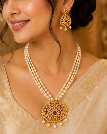 A woman wearing an ornate gold and pearl necklace with a large circular pendant and matching dangling earrings, styled with a beige blouse and draped saree in a warm indoor setting.