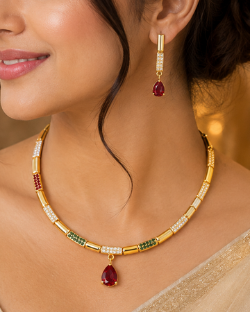 A woman wearing a gold-plated necklace and matching drop earrings with red and green stone accents, styled with a beige saree in a warm, elegant indoor setting.