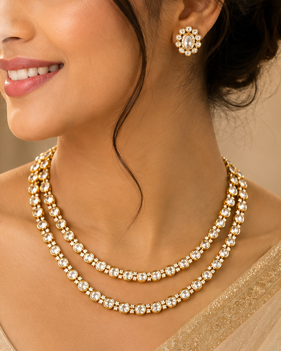 A woman wearing a gold double-strand necklace with clear stones and a matching stud earring, dressed in a beige traditional outfit against a softly blurred warm indoor background.