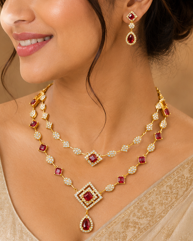A woman wearing a gold-toned traditional necklace and matching drop earrings with red gemstones, styled with a beige saree in a warm, elegant indoor setting.