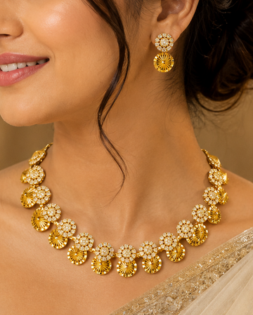 A woman wearing a gold-toned traditional necklace and matching drop earrings, paired with a light beige embroidered sari, shown in a warm indoor setting.