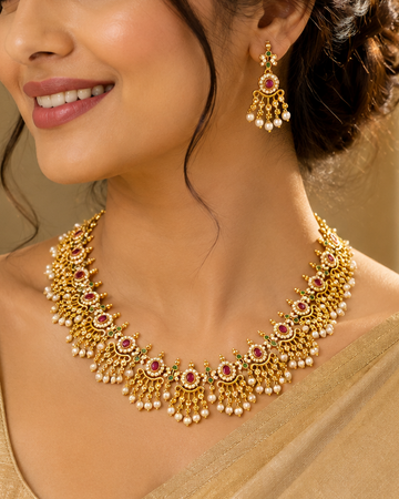 A woman wearing a gold-toned traditional necklace and matching drop earrings with red stones and white pearl-like accents, styled with a beige satin blouse in an indoor setting.
