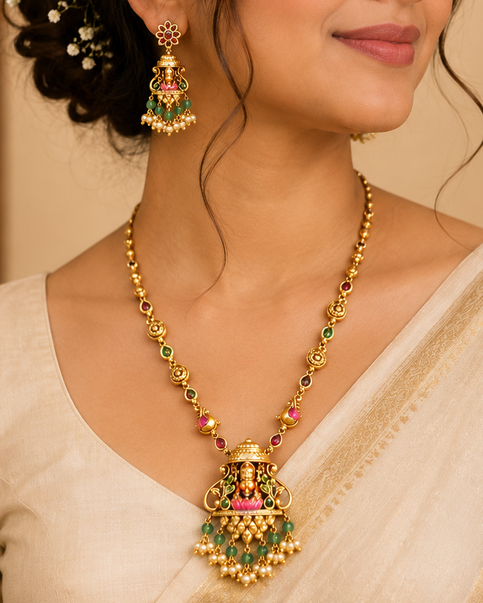 A woman wearing a traditional Indian gold necklace with a detailed pendant and matching earrings, styled with a cream-colored saree and hair accessory against a warm, softly blurred background.