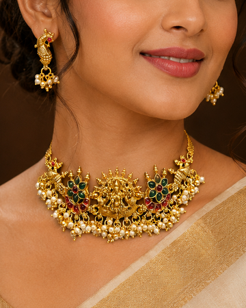 A woman wearing an ornate gold-toned traditional necklace with green and red stone accents and matching earrings, styled with a beige saree in an elegant indoor setting.