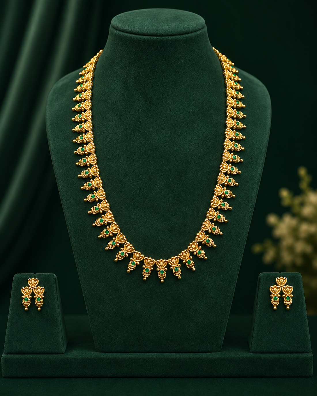 A woman wearing a gold necklace and matching gold earrings with green stone accents, styled with a cream saree against a dark green background.