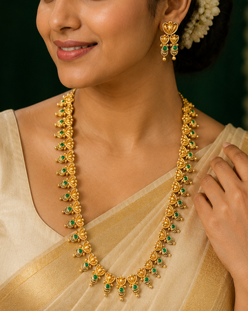 A woman wearing a gold necklace and matching gold earrings with green stone accents, styled with a cream saree against a dark green background.