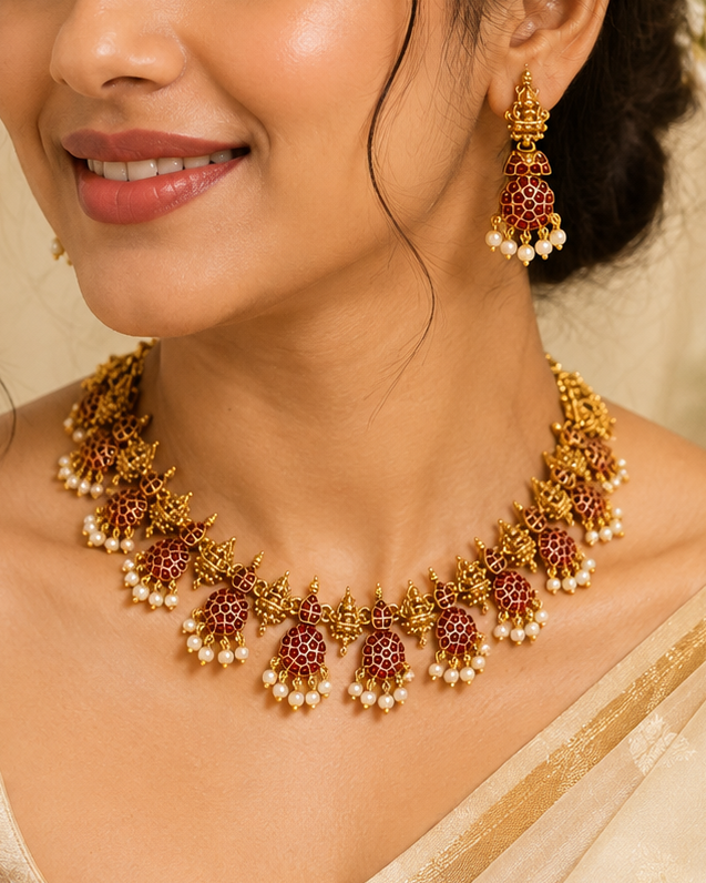 A woman wearing an ornate gold-toned traditional necklace with maroon floral motifs and white pearl-like beads, paired with matching dangling earrings, against a soft cream background.