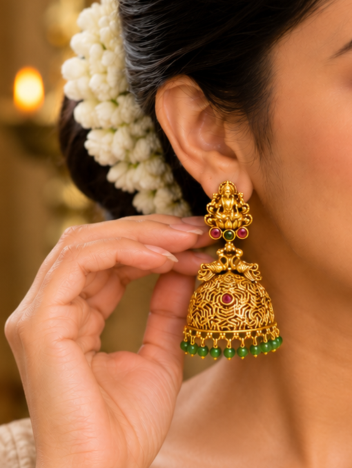 A woman wearing ornate gold jhumka earrings with green stone accents, shown in a close-up portrait. She is dressed in a light beige saree and has white flowers in her hair, set against a warm, softly blurred indoor background.