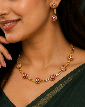 A woman wearing a gold-toned necklace and matching drop earrings with pink stone accents, styled with a dark green sari against a dark green draped background.