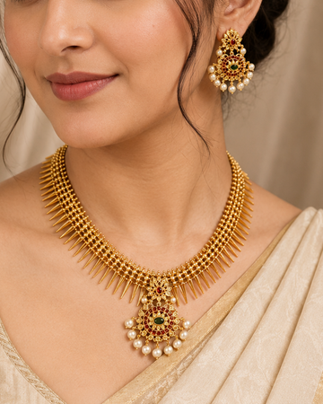 A woman wearing an ornate gold necklace and matching earrings with pearl-like white stones and small green accents, styled with a beige sari against a soft beige draped backdrop.