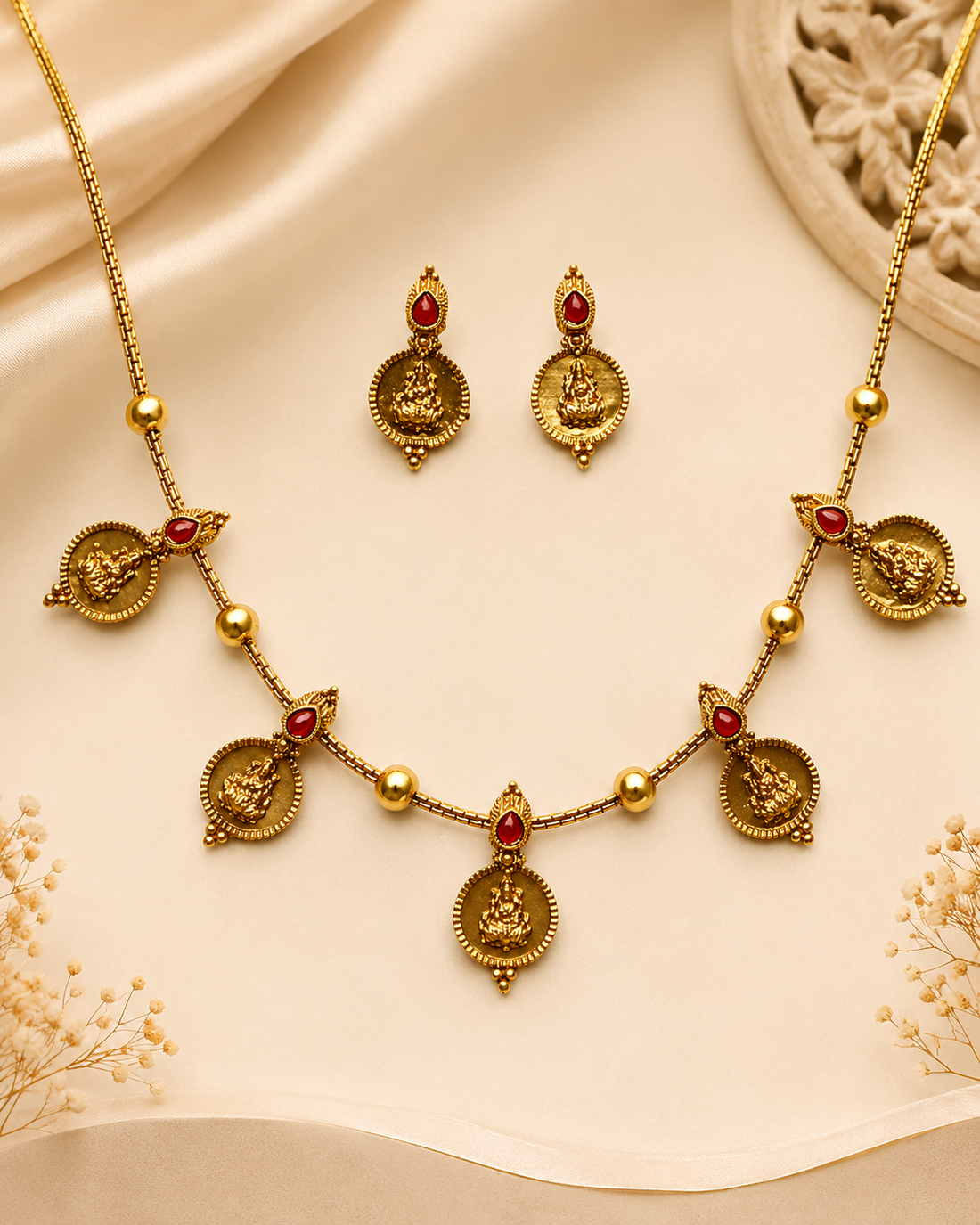 A woman wearing a gold necklace and matching earring with small red stone accents, styled with a cream saree and white hair flowers against a warm beige background.