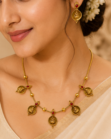 A woman wearing a gold necklace and matching earring with small red stone accents, styled with a cream saree and white hair flowers against a warm beige background.