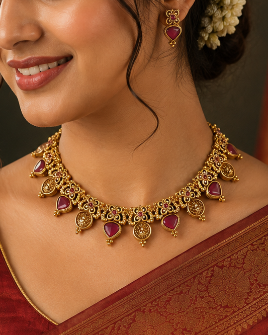 A woman wearing a red and gold brocade saree with a matching gold necklace and earrings adorned with pink stones and white flowers in her hair, set against a dark background with a red patterned curtain.