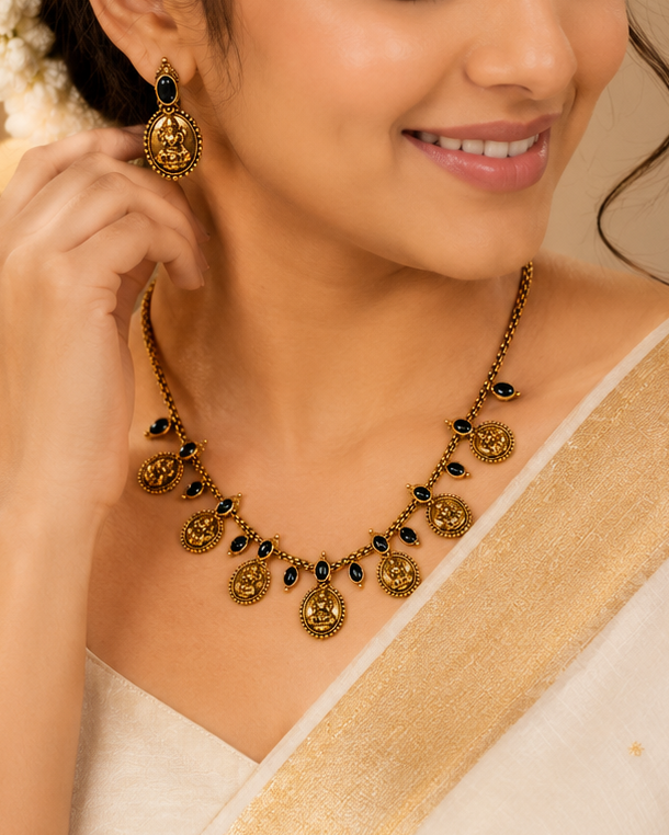A woman wearing a cream and gold saree with a matching gold necklace and earrings, adorned with a white floral hair accessory, in a warm, softly lit indoor setting.