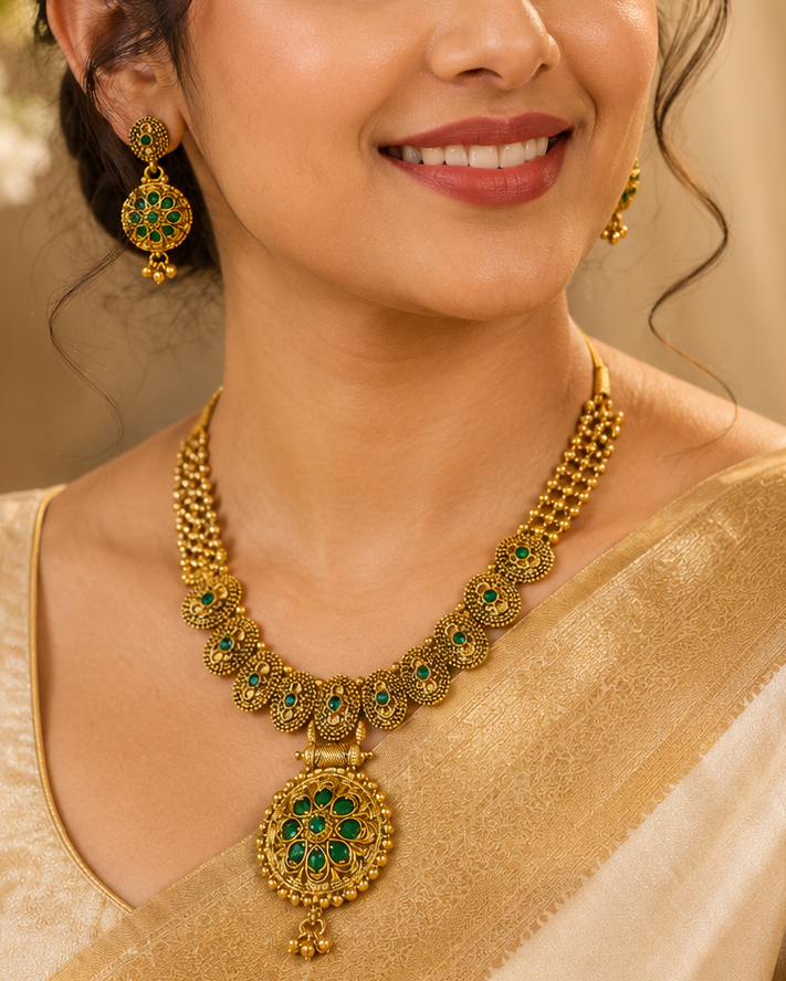 A woman wearing a gold-toned traditional necklace with green stone accents and matching dangling earrings, styled with a cream-colored saree against a soft beige background.