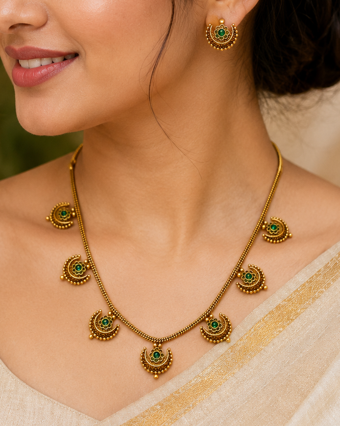 A woman wearing a gold-toned necklace and matching earrings with green stone accents, paired with a beige sari. The jewelry is shown in a close-up portrait against a softly blurred warm neutral background.