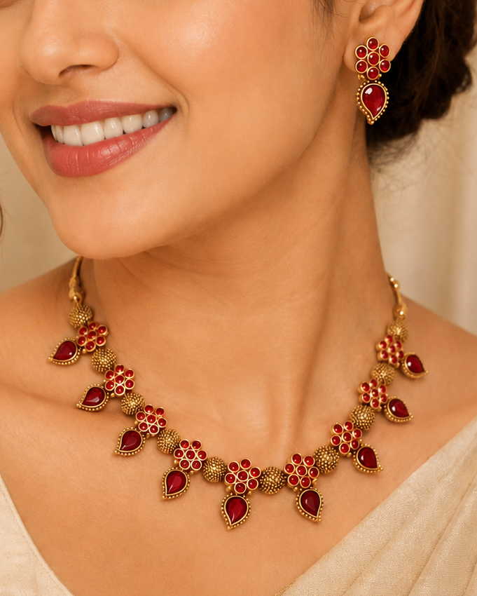 A woman wearing a gold and red traditional Indian jewelry set, featuring a necklace and matching earrings, styled with a beige sari.