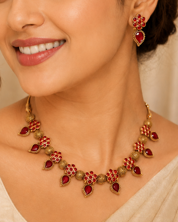 A woman wearing a gold and red traditional Indian jewelry set, featuring a necklace and matching earrings, styled with a beige sari.