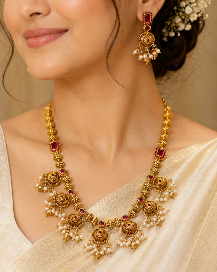 A woman wearing an ornate gold-toned traditional necklace with red stones and pearl-like accents, matching drop earrings, and a cream sari against a softly blurred beige background.