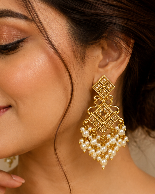 A woman wearing a large, ornate gold-toned dangle earring with white pearl-like beads, shown in a close-up portrait. She is wearing a beige embroidered sari, and the background is a softly blurred warm indoor setting.