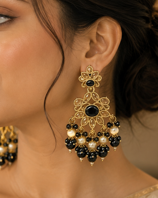 A woman in profile wearing ornate gold-toned traditional earrings with black and white bead drops, paired with a cream-colored embroidered saree blouse.