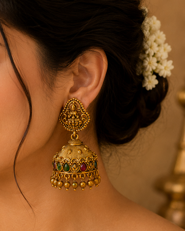 A woman in a beige traditional outfit wearing a large ornate gold jhumka earring with red, green, and blue stone accents, photographed in warm indoor lighting.