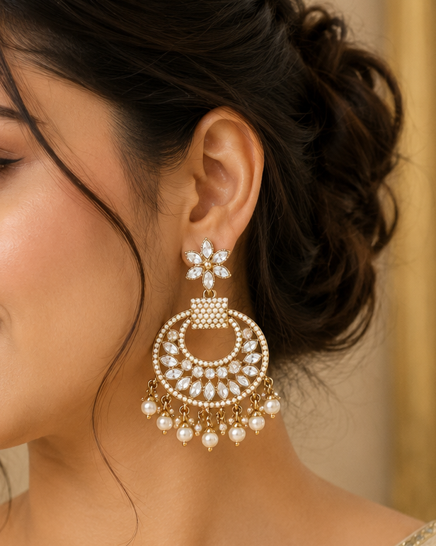 A woman wearing a large gold-toned huggie chandelier earring adorned with white faux pearls and crystal-like details, shown in a close-up portrait.
