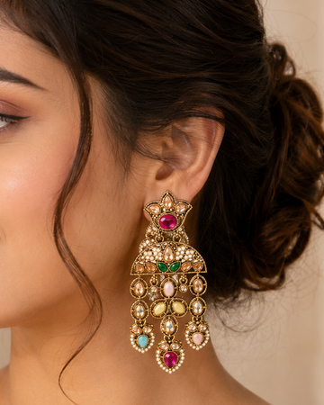 A woman wearing a pair of ornate gold-toned chandelier earrings with red, green, and blue stones, shown in a close-up portrait.
