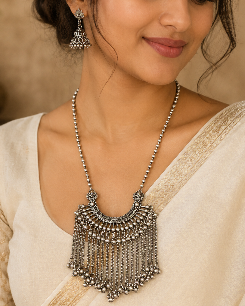 A woman wearing a traditional silver-toned Indian necklace with a large, ornate fringed pendant and matching jhumka earrings, styled with a cream-colored saree.