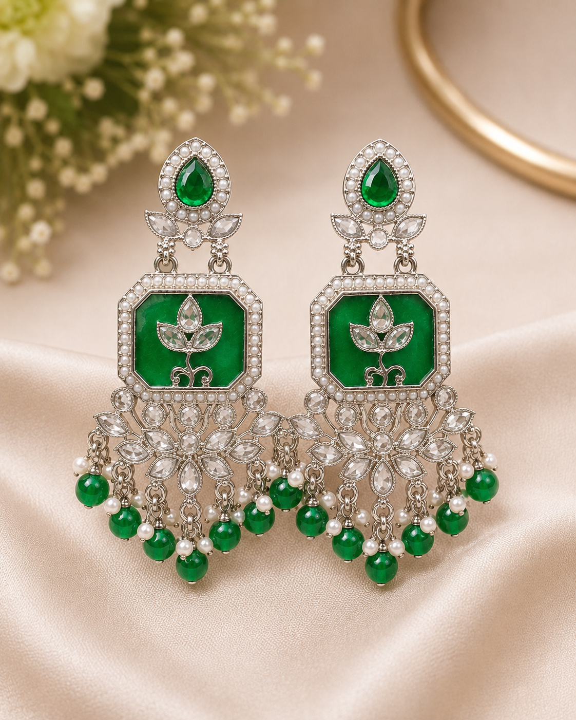A pair of ornate silver-toned chandelier earrings with emerald green inlay panels, clear pavé stones, and dangling green bead drops, displayed on a beige satin fabric background.