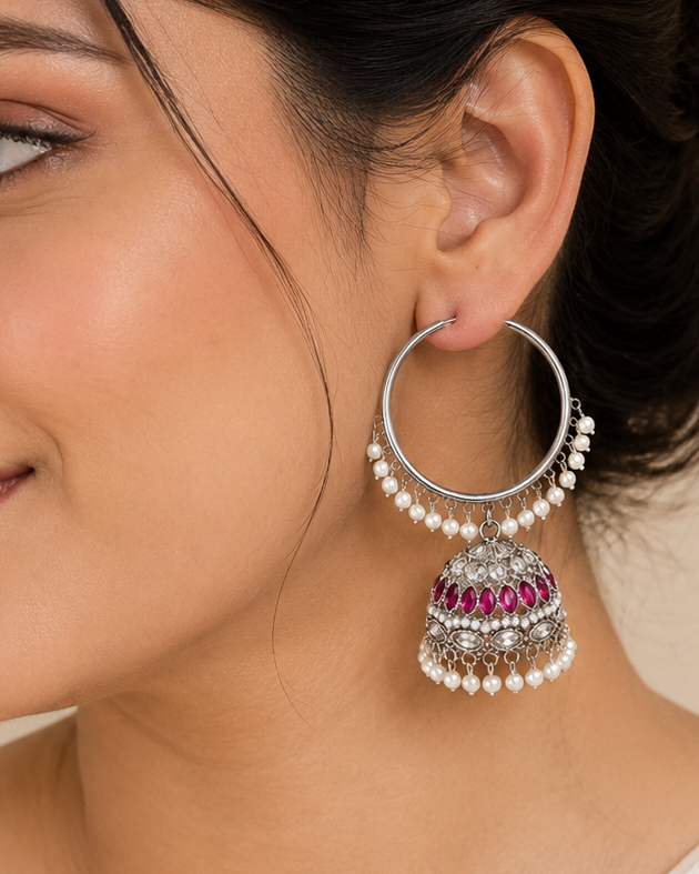 A woman wearing a pair of silver-toned traditional Indian jhumka earrings with white pearl-like beads and pink gemstone detailing, shown in a close-up profile shot.