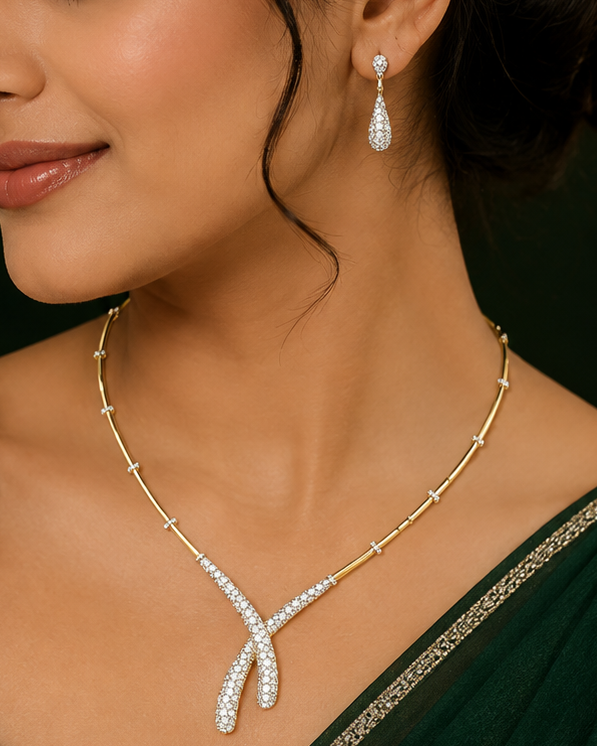 A woman wearing a gold Y-shaped necklace and matching teardrop earring, both adorned with sparkling clear stones. She is wearing a dark green sari in a warmly lit indoor setting.