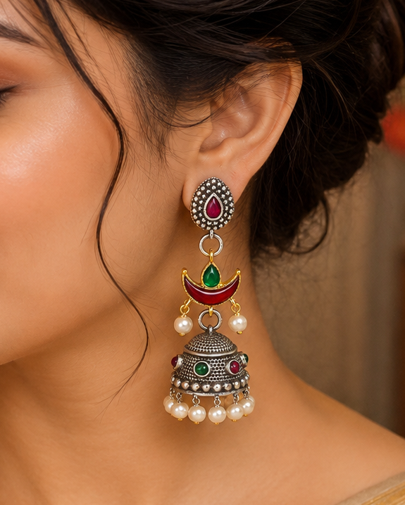 A woman wearing a traditional Indian jhumka earring with silver-toned metal, red and green stones, and white faux pearls, paired with a beige saree.