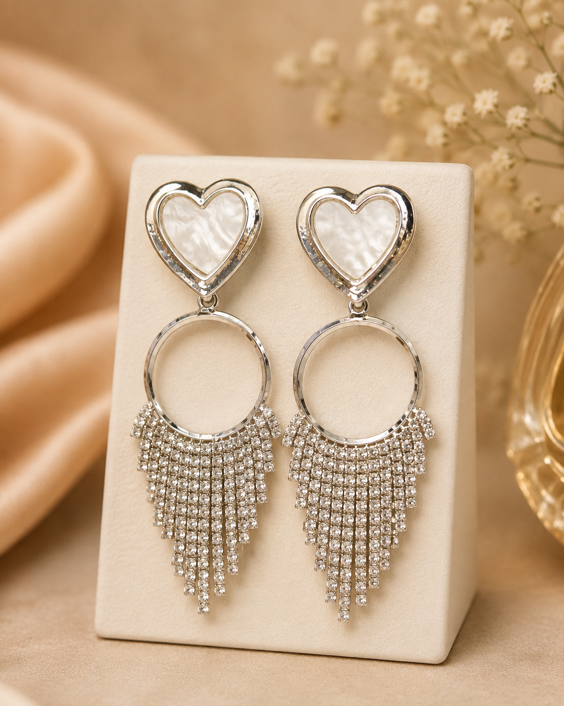 A close-up of a woman wearing a pair of silver-toned statement earrings with a heart-shaped crystal top and dangling crystal fringe, against a warm beige background.